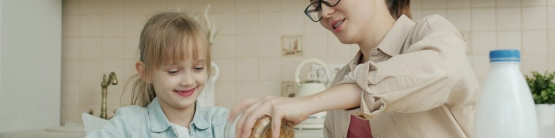 Young woman is making breakfast and talking to cute little daughter at home in modern cozy kitchen. Food, motherhood and apartment concept.